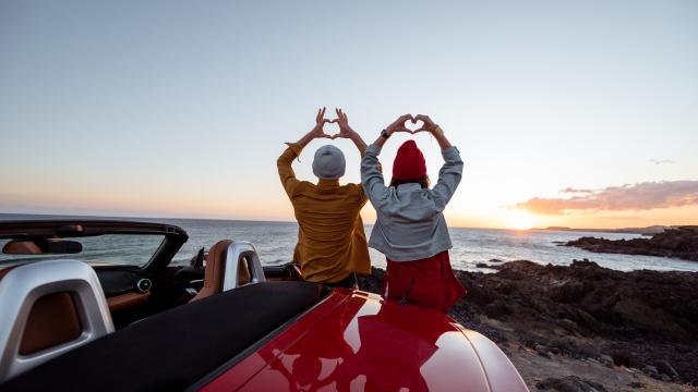 Couple enjoying beautiful views on the ocean, standing together near the car on the rocky coast, showing with hands heart shape. Carefree lifestyle, love and travel concept